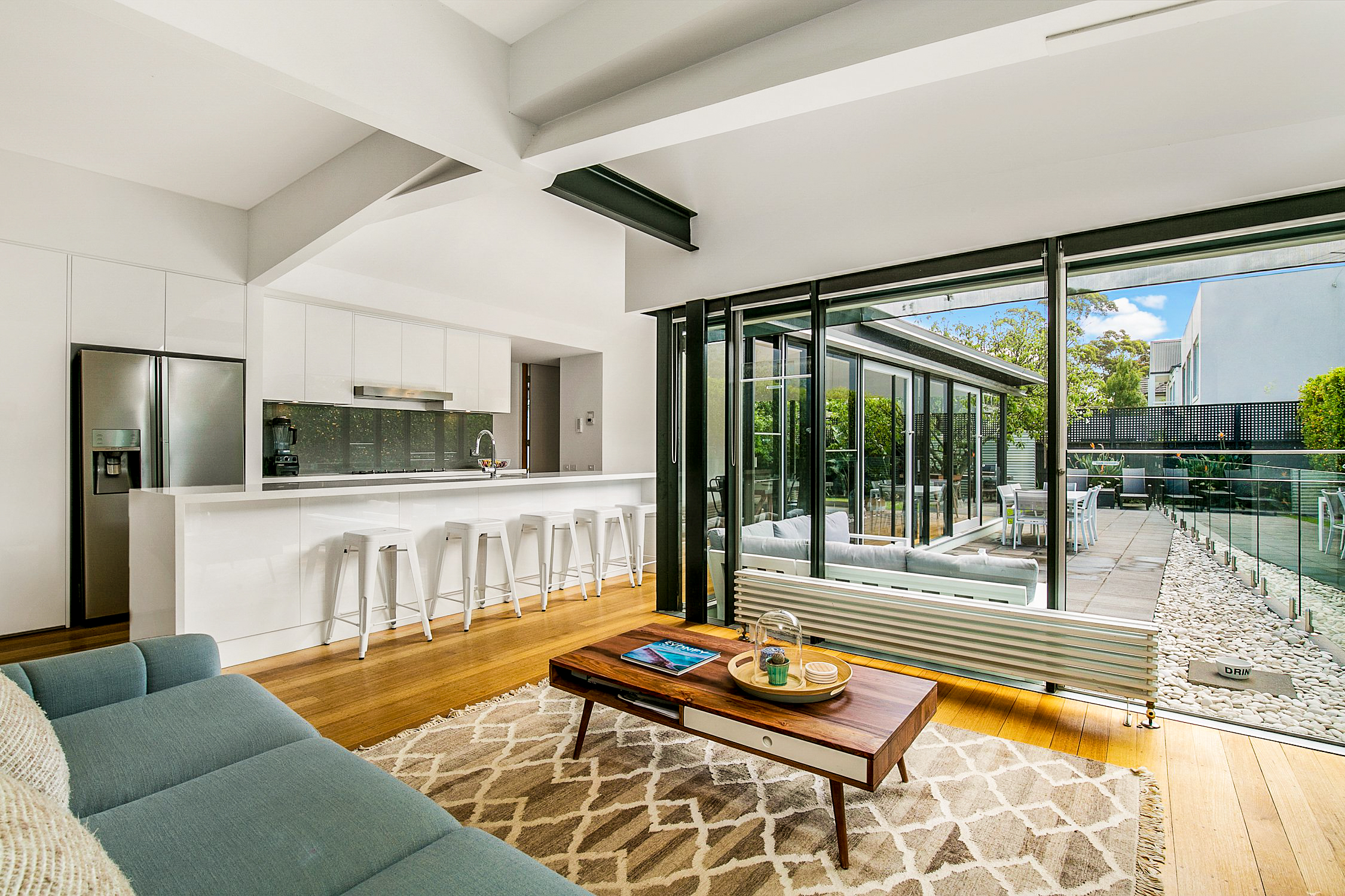 Bright modern living room with blue sofa, wooden coffee table, kitchen island, and glass doors to outdoor patio.