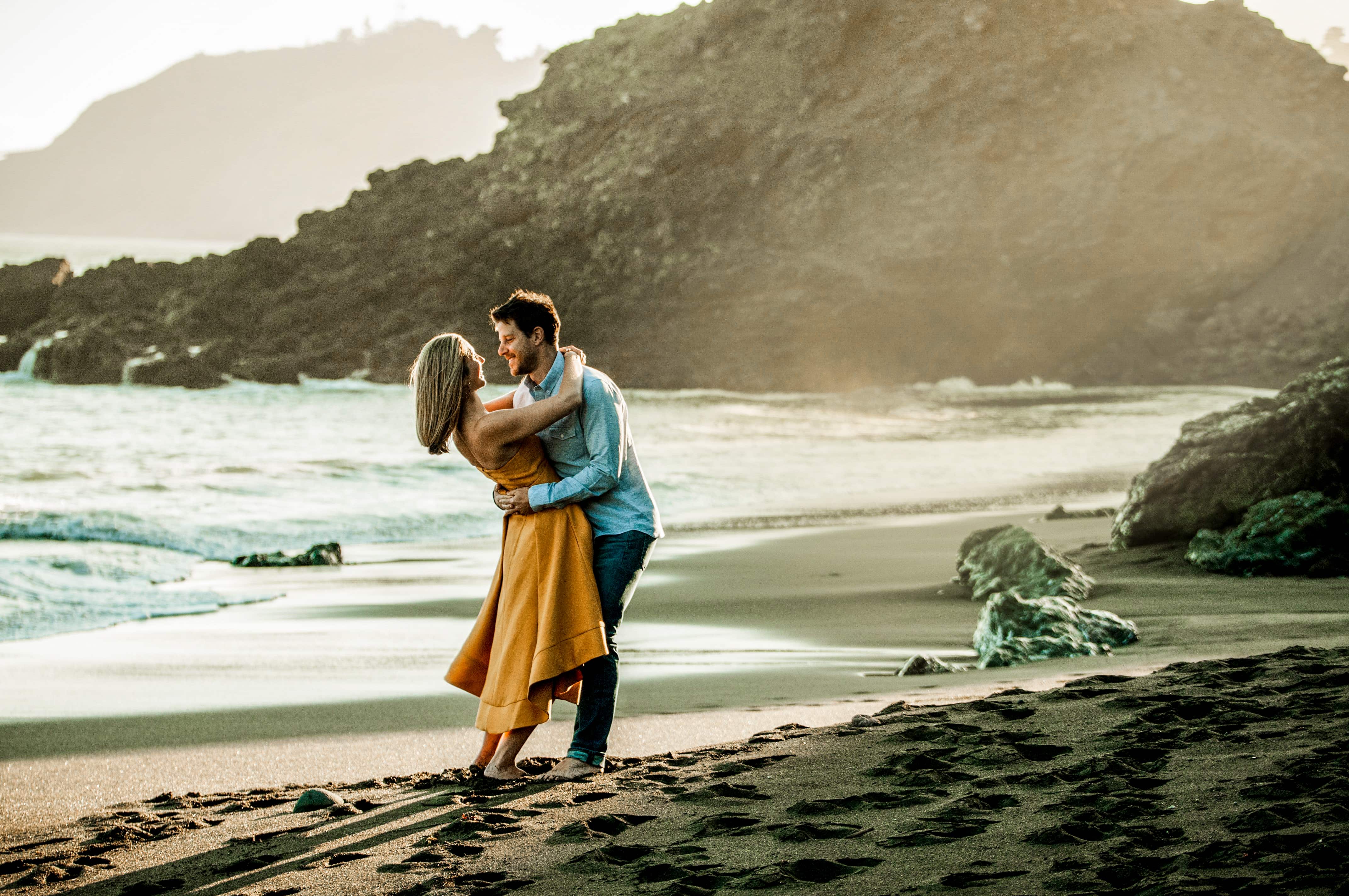 Couple embracing on a sandy beach with rocky cliffs and ocean waves in the background.