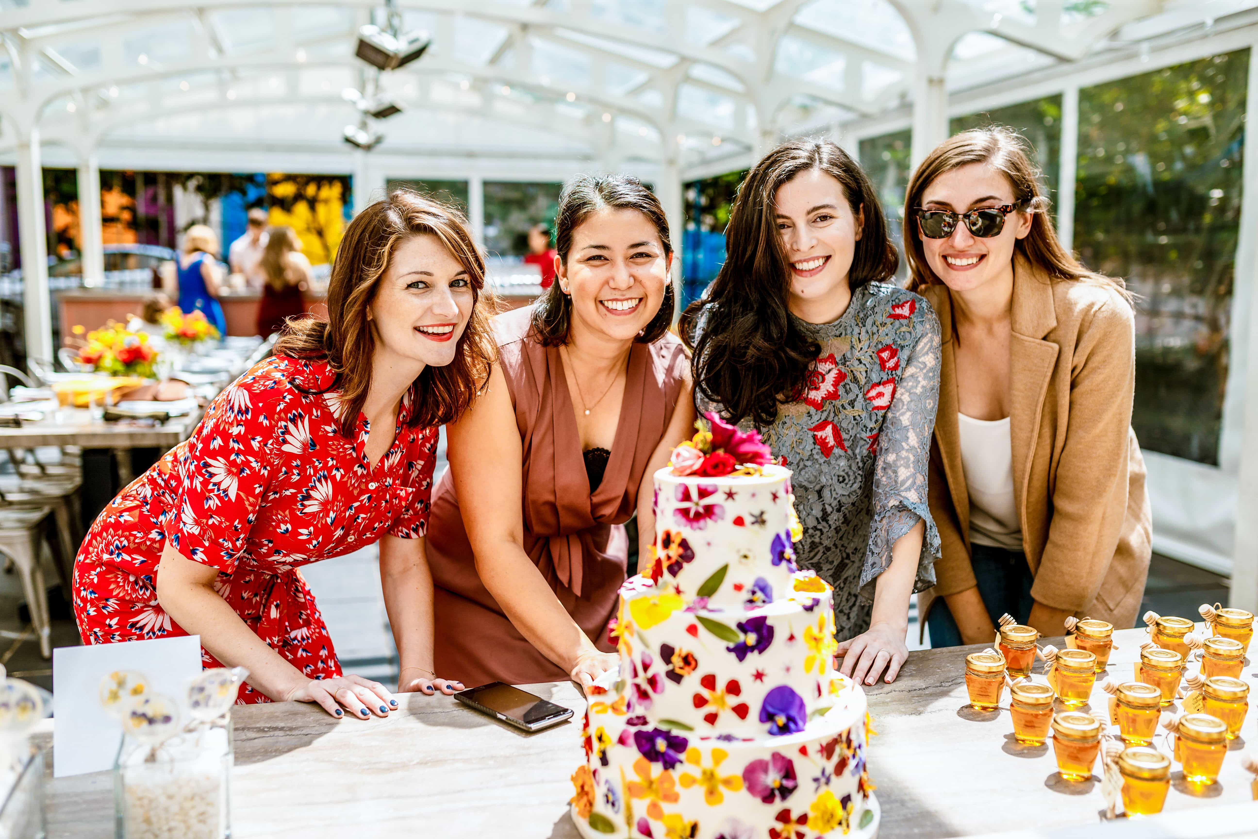 Four smiling women leaning over a table with a three-tier floral cake and small jars of honey.