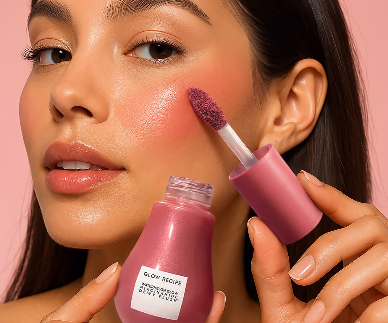 Close-up of a woman applying pink cream blush with applicator to her cheek, holding a Glow Recipe Watermelon Glow Niacinamide Dewy Flush bottle.