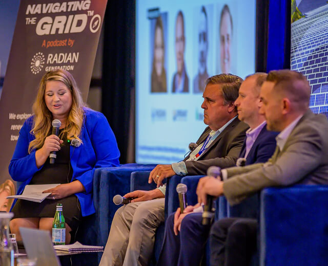 Four panelists engaged in a corporate discussion, with one woman speaking into a microphone and holding notes.