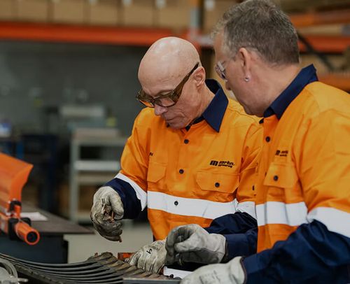 Two male workers in orange high-visibility shirts inspecting and working with metal parts in an industrial setting.