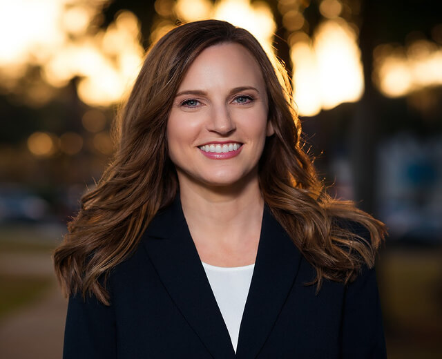Smiling woman with long brown hair wearing a dark blazer and white top, outdoors at sunset.