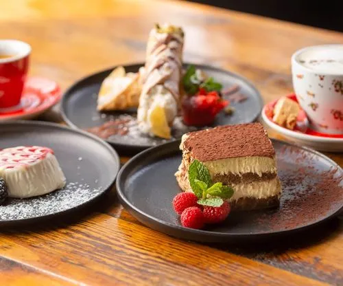 Tiramisu dessert garnished with raspberries and mint on a black plate, with other desserts and a cup of coffee on a wooden table.