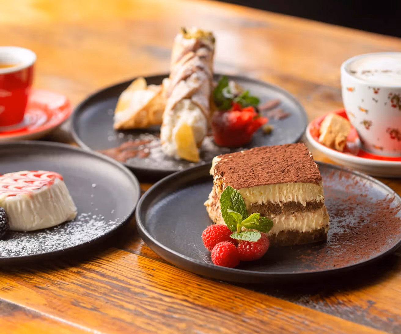 Tiramisu dessert garnished with raspberries and mint on a black plate, with other desserts and a cup of coffee on a wooden table.