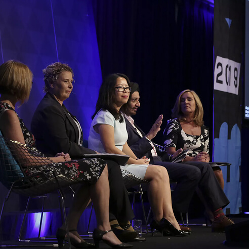 Five diverse professionals seated on stage panel during a business event with a 2019 banner in the background.