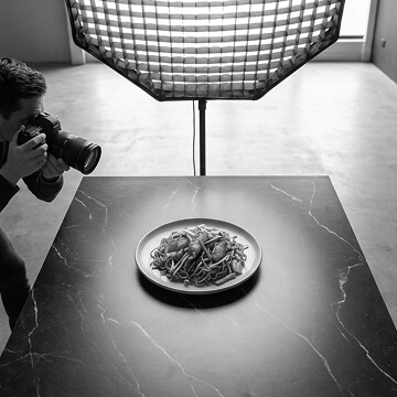 Photographer capturing a plate of food on a dark marble table with a large softbox light above.