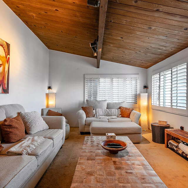 Cozy living room with vaulted wood ceiling, beige sofas with brown pillows, a wooden coffee table, and soft lighting.