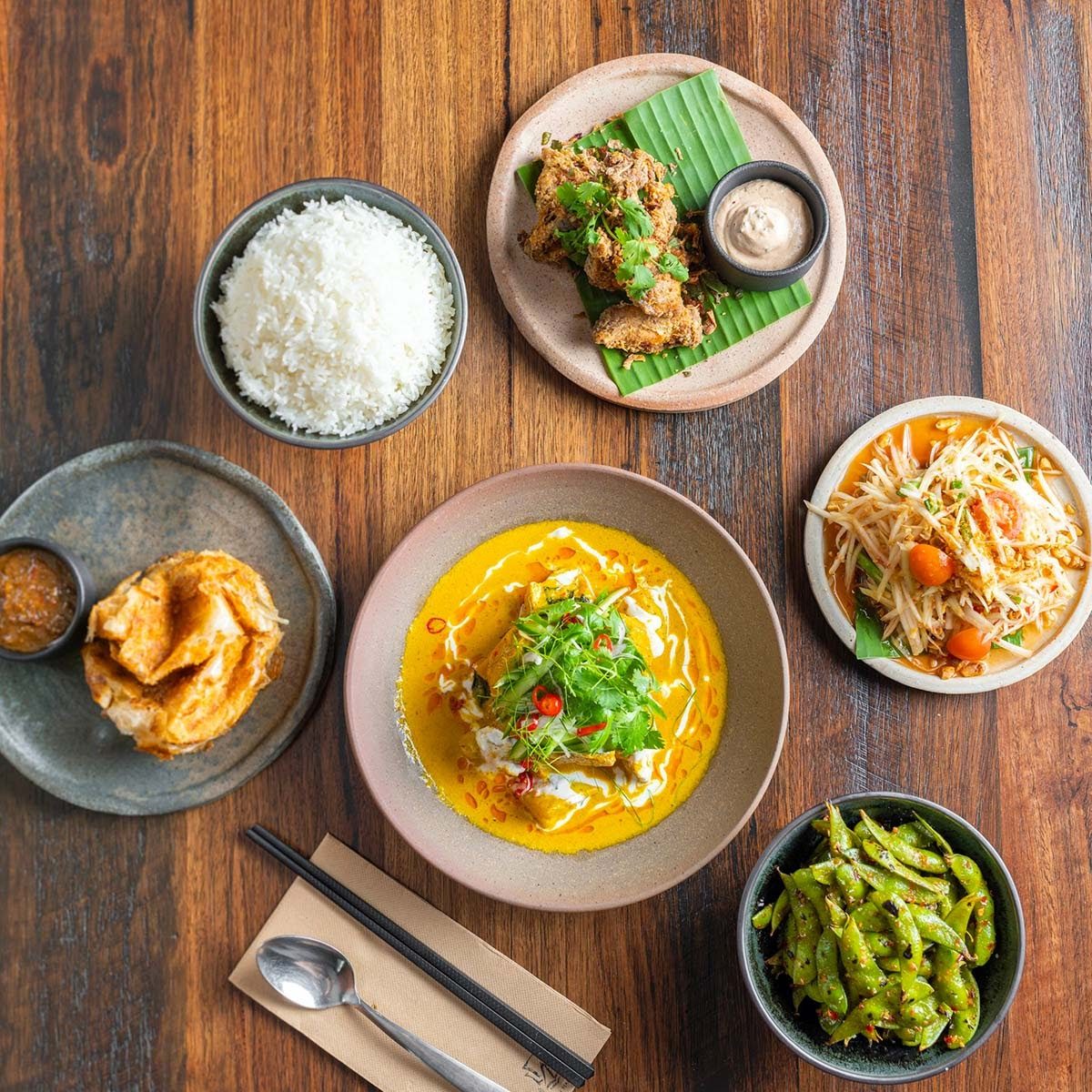 Top view of Thai dishes including curry, rice, fried chicken, salad, and green beans on wooden table.
