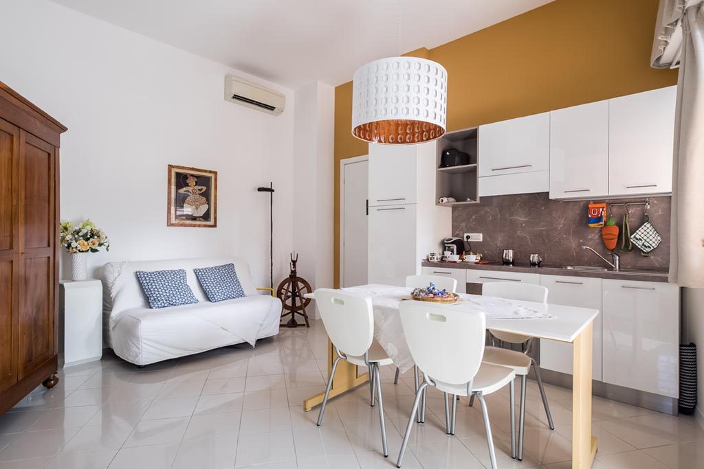 Modern kitchen and dining area with white cabinets, table, chairs, sofa, and wooden wardrobe.