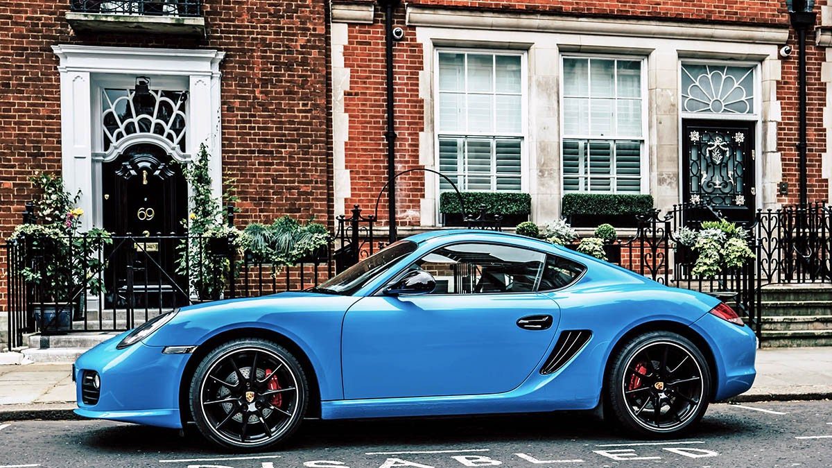 Blue Porsche Cayman parked on a city street with red brick buildings and black doors in background.