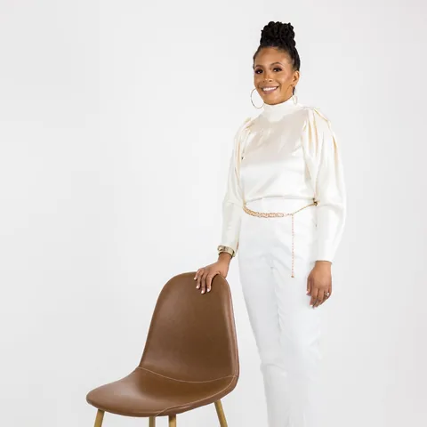 Woman in a white satin outfit posing beside a tan chair in a bright studio