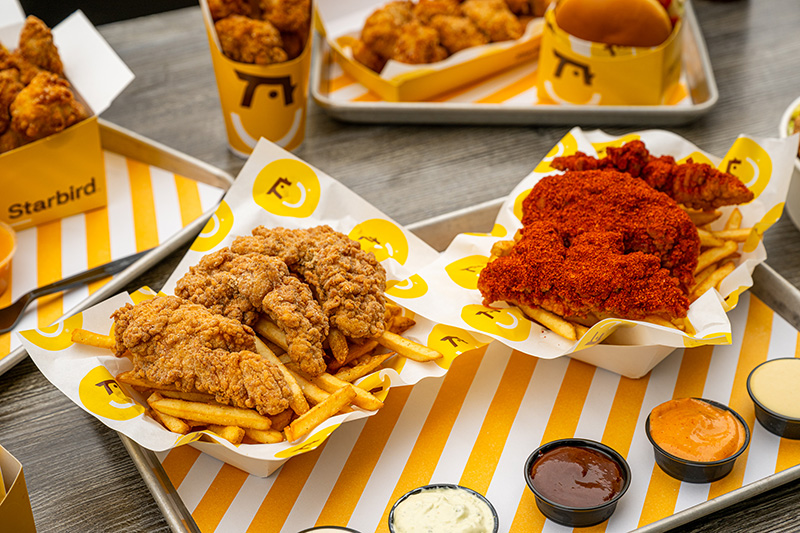 Trays with crispy fried chicken tenders and French fries served with multiple dipping sauces on a wooden table.