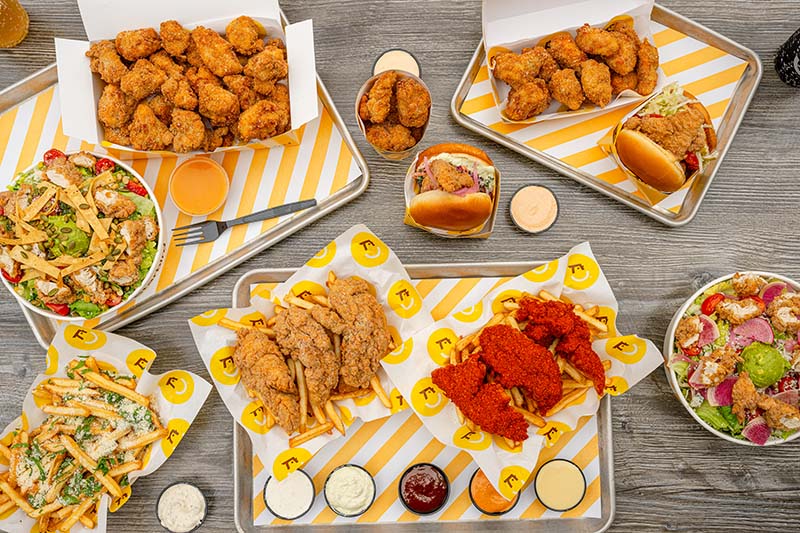 Variety of fried chicken tenders, two chicken sandwiches, loaded fries, salads, and dipping sauces arranged on trays on a wooden table.