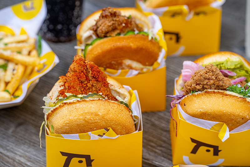 Three crispy fried chicken sandwiches in yellow branded boxes on a wooden table with fries and a drink in the background.