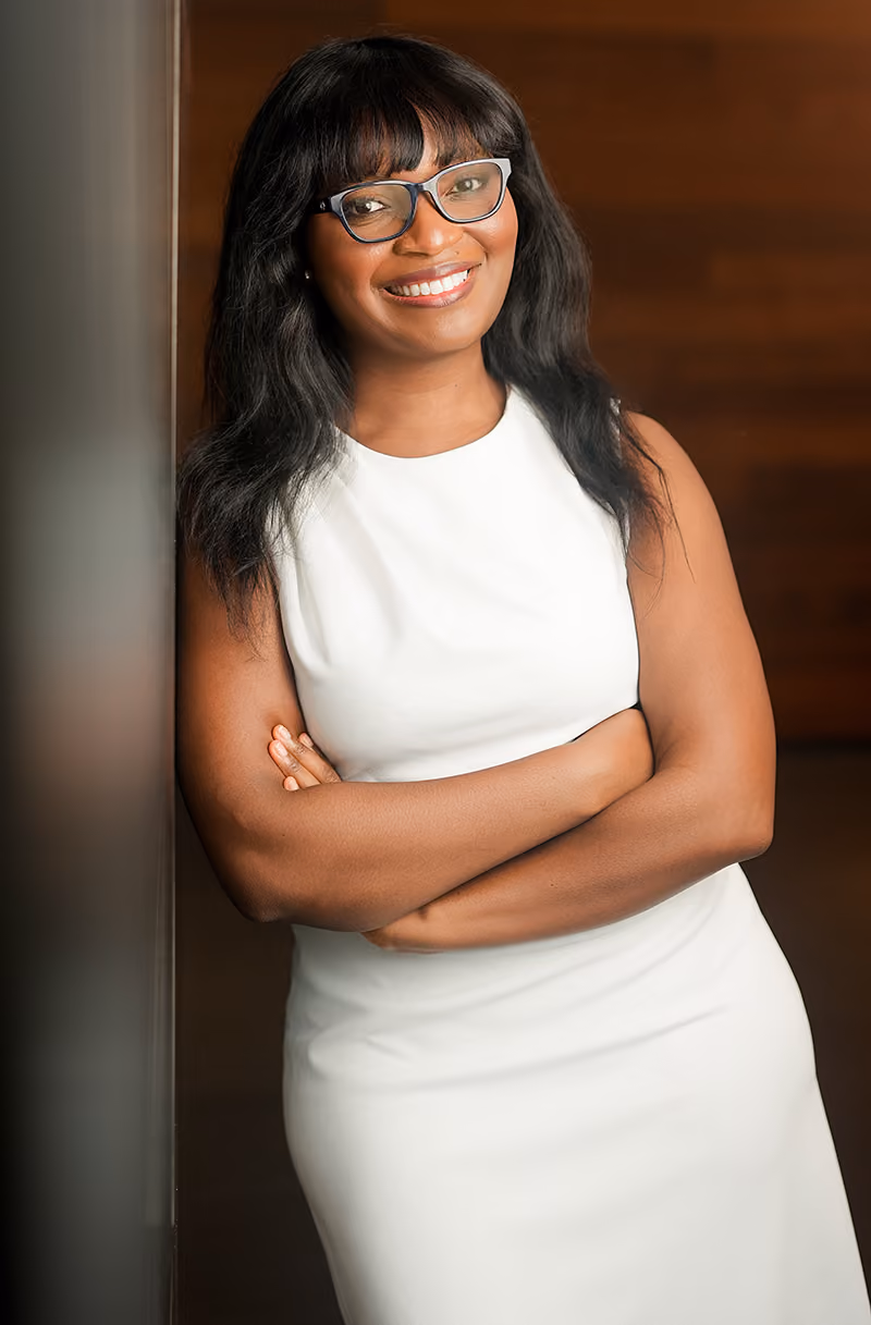 Smiling woman with long dark hair and glasses, wearing a white dress and leaning on a wall with folded arms