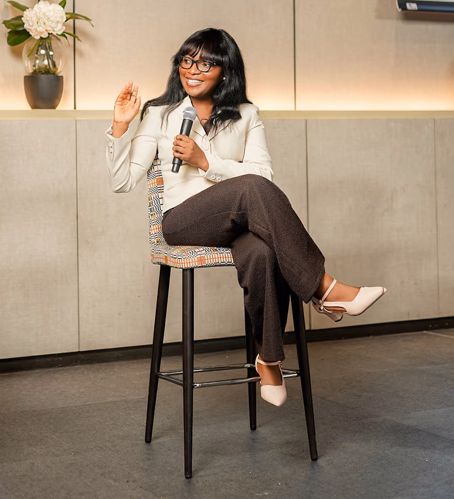 A woman with glasses, wearing a white blazer and dark pants, sits cross-legged on a tall stool holding a microphone and gesturing with her left hand, in a modern indoor setting with a flower vase on a shelf behind her.