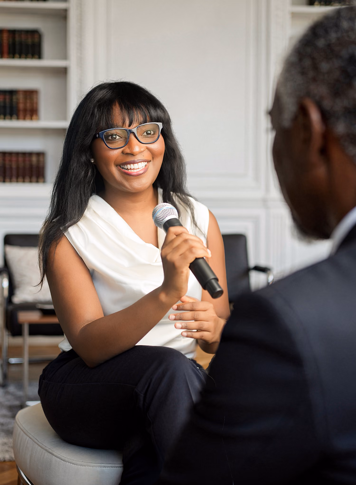 Smiling woman with glasses holding a microphone, speaking to a man in a suit in a room with bookshelves.