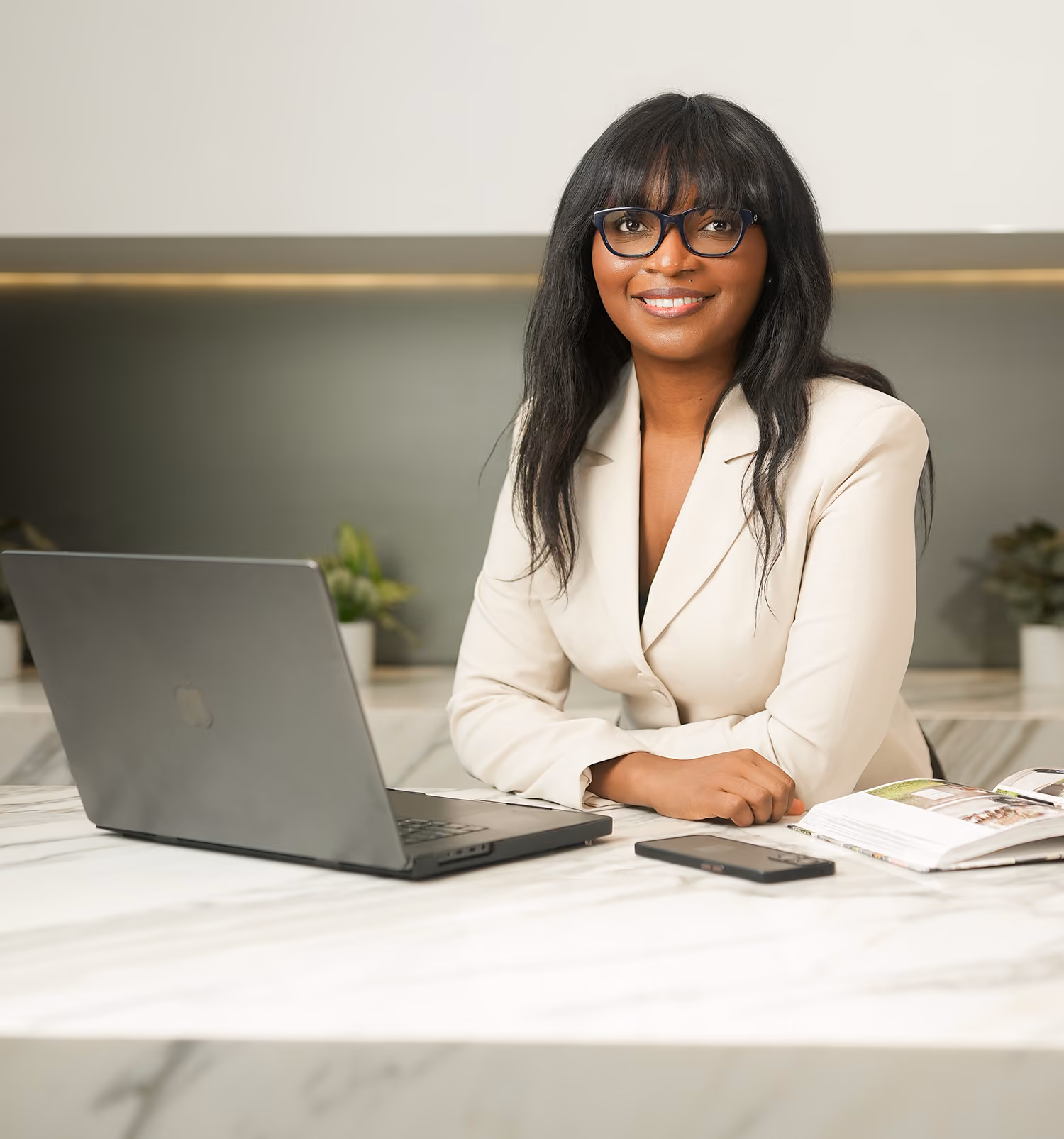 Smiling woman with long black hair and glasses seated at a marble table with a laptop, smartphone, and open book in a modern office setting.