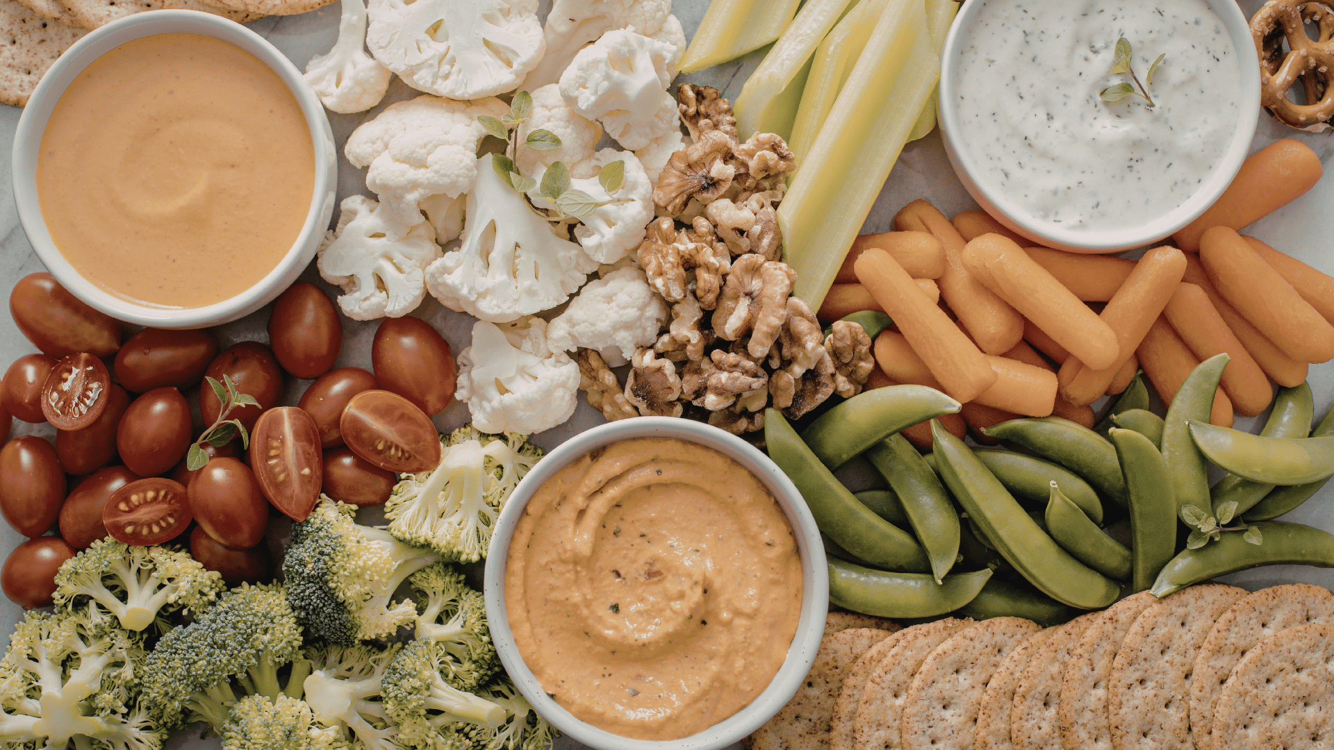 An overhead view of an array of veggies, crackers, nuts, and dips