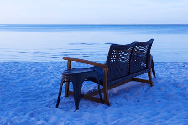 Black woven bench with wooden armrests and a black metal stool on white sandy beach by calm ocean during twilight.
