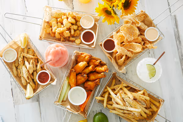 Five baskets with fried chicken wings, fries, fried fish, crispy pork rinds, and fried potatoes with dipping sauces and two beverages on a white table with sunflowers.