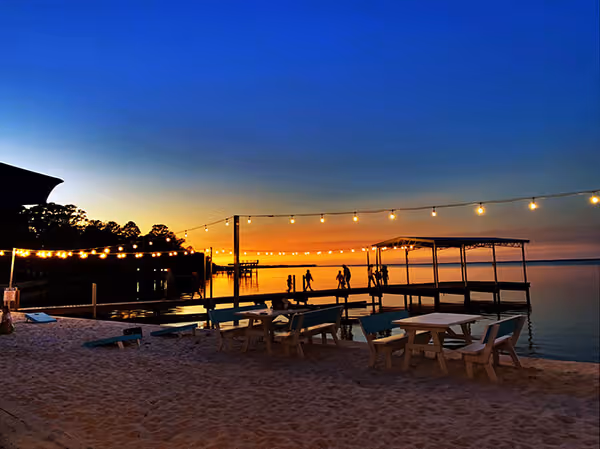 Wooden pier with people silhouetted at sunset over calm water, beach chairs and string lights in foreground.