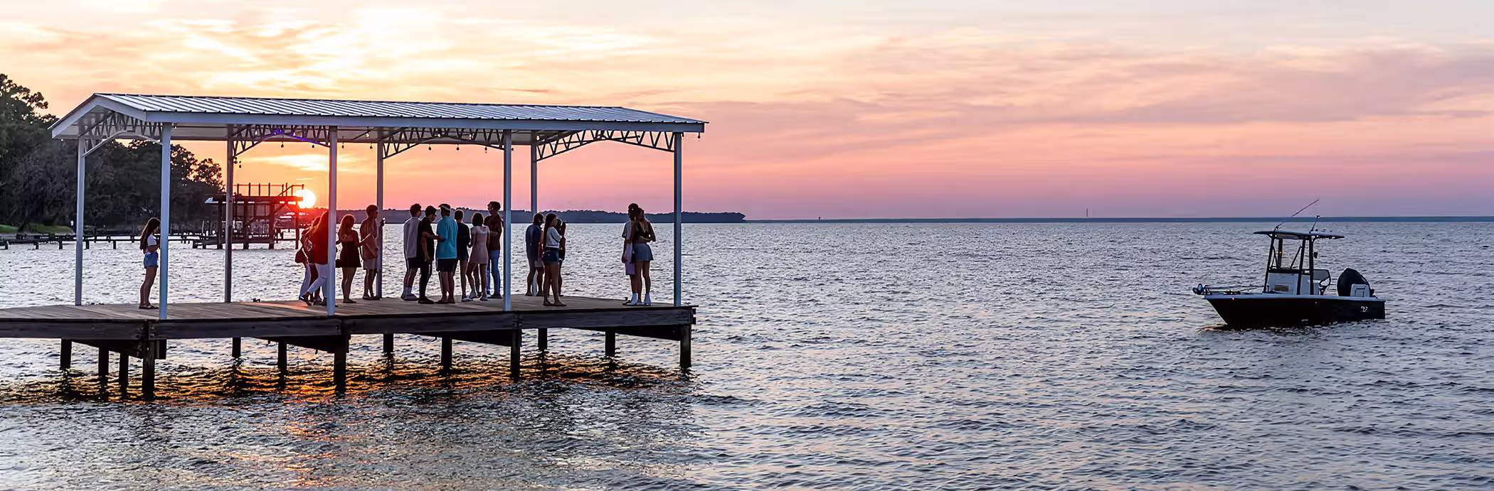 Group of people gathered on a covered wooden pier at sunset with a boat floating nearby on calm water.