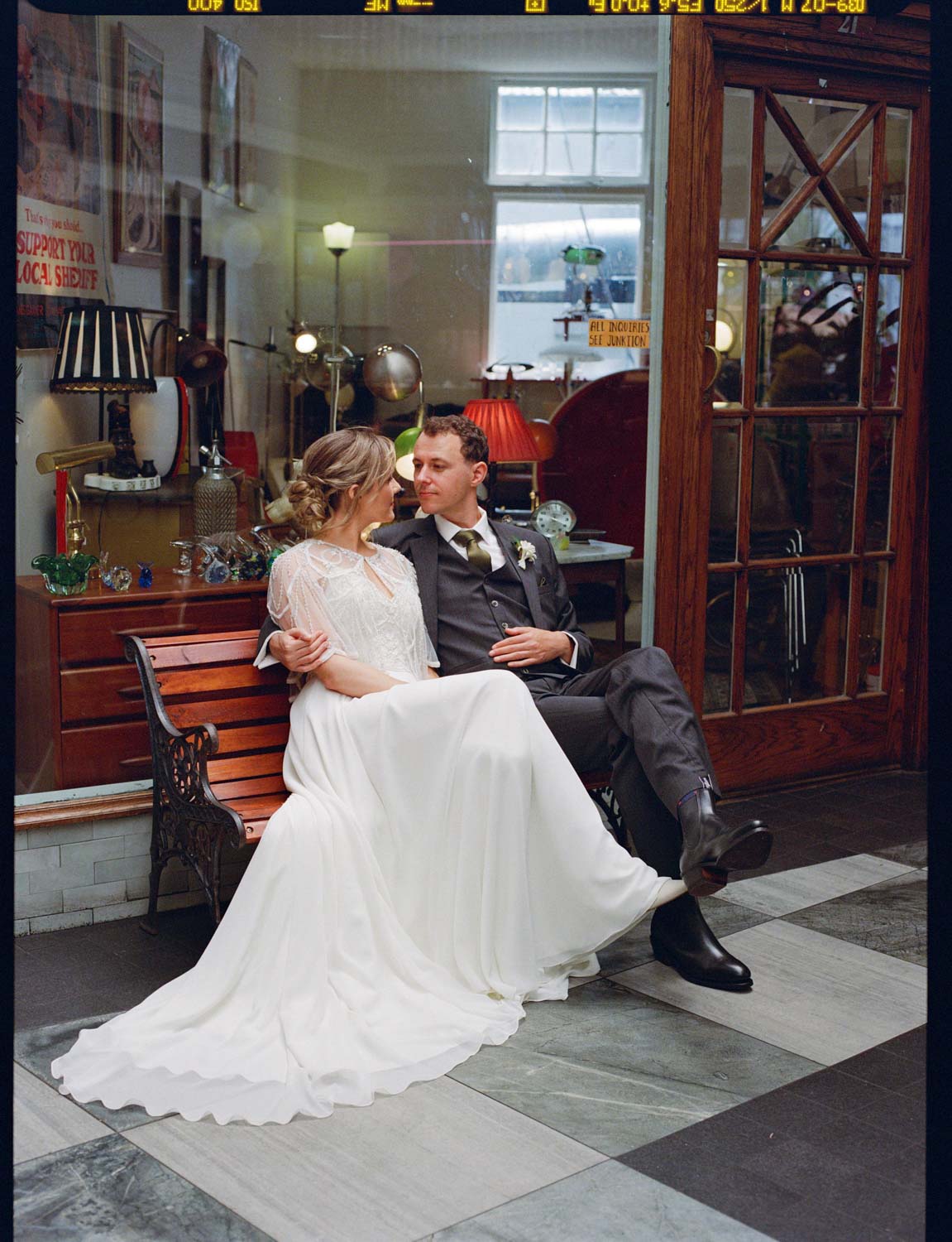 Analog medium format photo of cool bride and groom sitting on a bench in front of a second hand store at the Arcade on Karangahape Road Auckland
