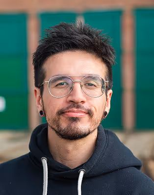 Portrait of a man with glasses, a short beard, and black hoodie, against a blurred outdoor background.