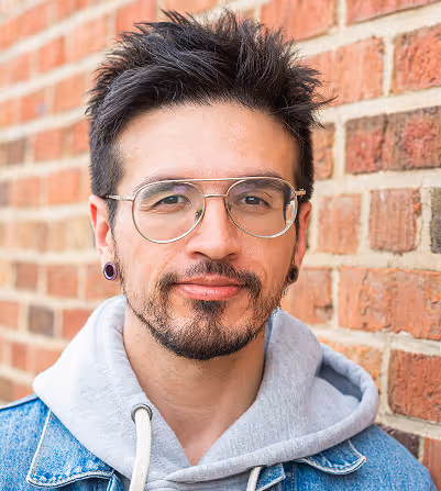 Man with short spiked hair, glasses, earrings, wearing a gray hoodie and denim jacket standing against a brick wall.