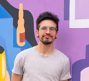 Smiling man with glasses and a light gray t-shirt standing in front of a colorful mural with abstract shapes.