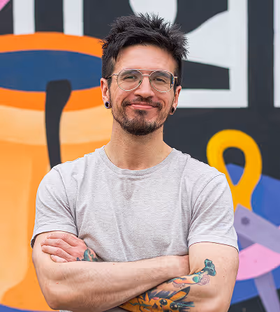 Man with glasses, tattoos, and earrings wearing a gray t-shirt, standing with arms crossed in front of a colorful mural.