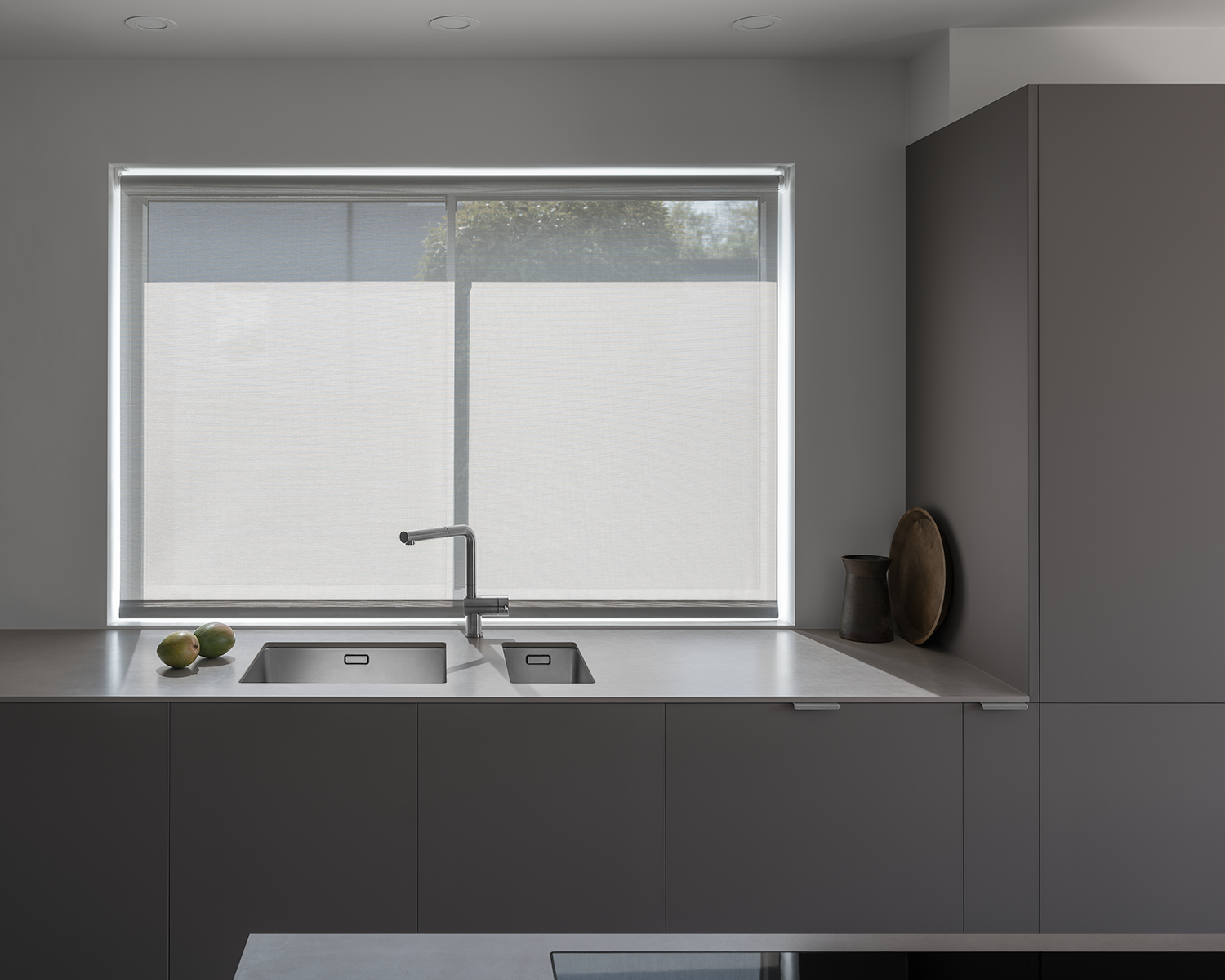 Japanese, minimal kitchen with white walls, light grey blind, grey brown cabinets, sintered stone worktop, stainless steel tap, wooden tray and mangos