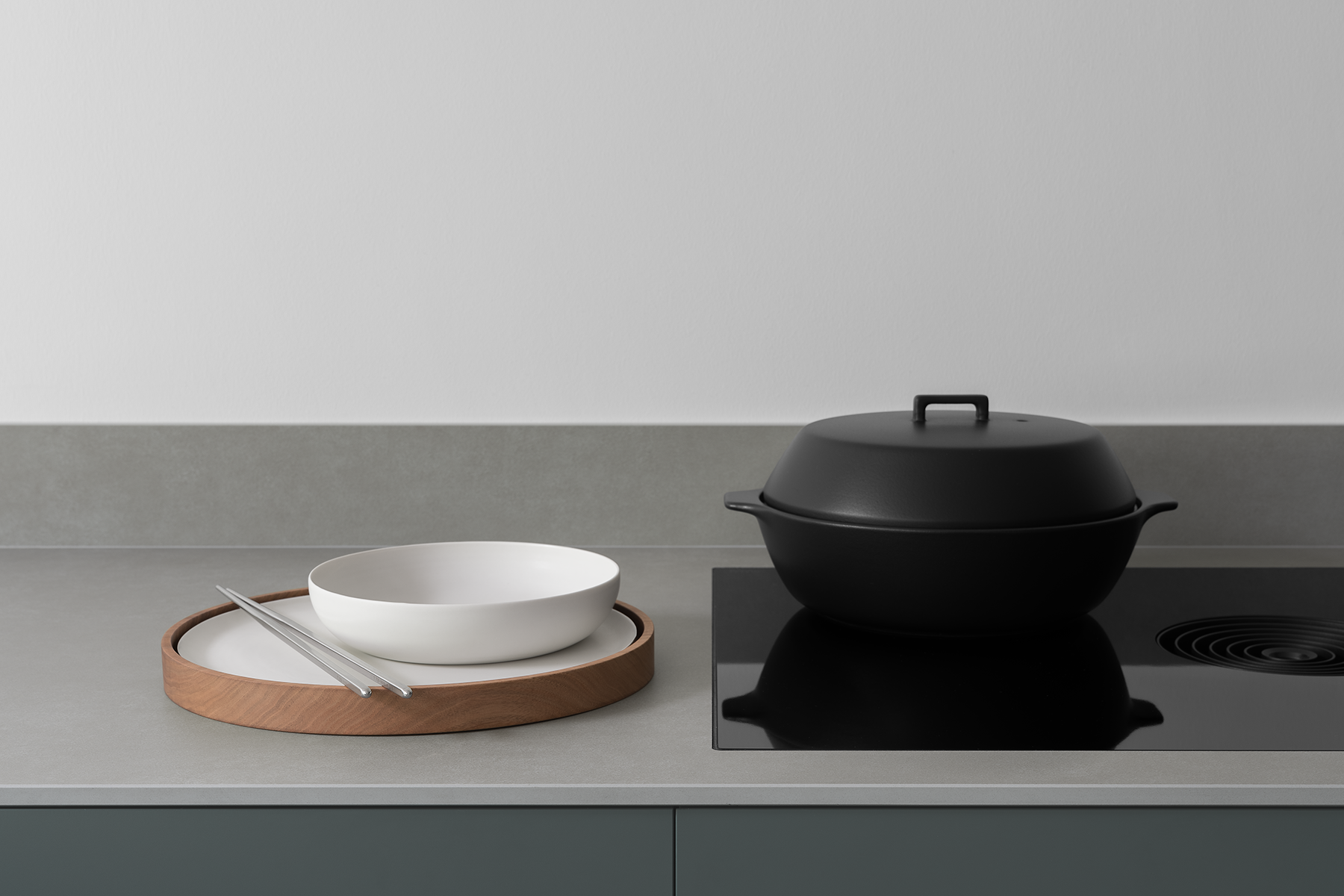 Modern slim textured stone kitchen work surface with black induction hob with integral extractor. Japanese black ceramic donabe, white porcelain bowl and plate on round wooden tray with stainless steel chopsticks