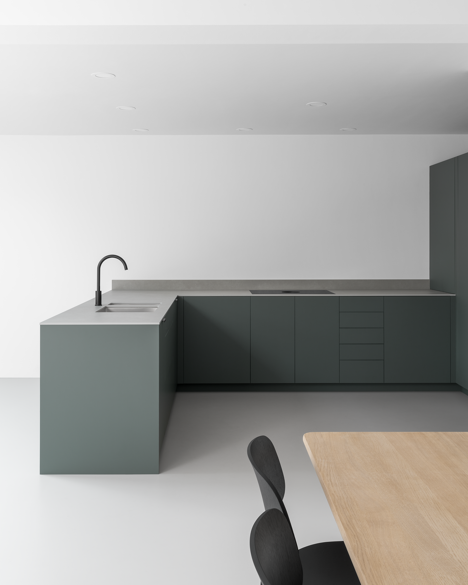 Japanese style minimal modern white kitchen dining room with grey resin floor. Dark green-grey cabinets, textured grey slim work surface, black tap, Danish oak dining table with modern black chairs.