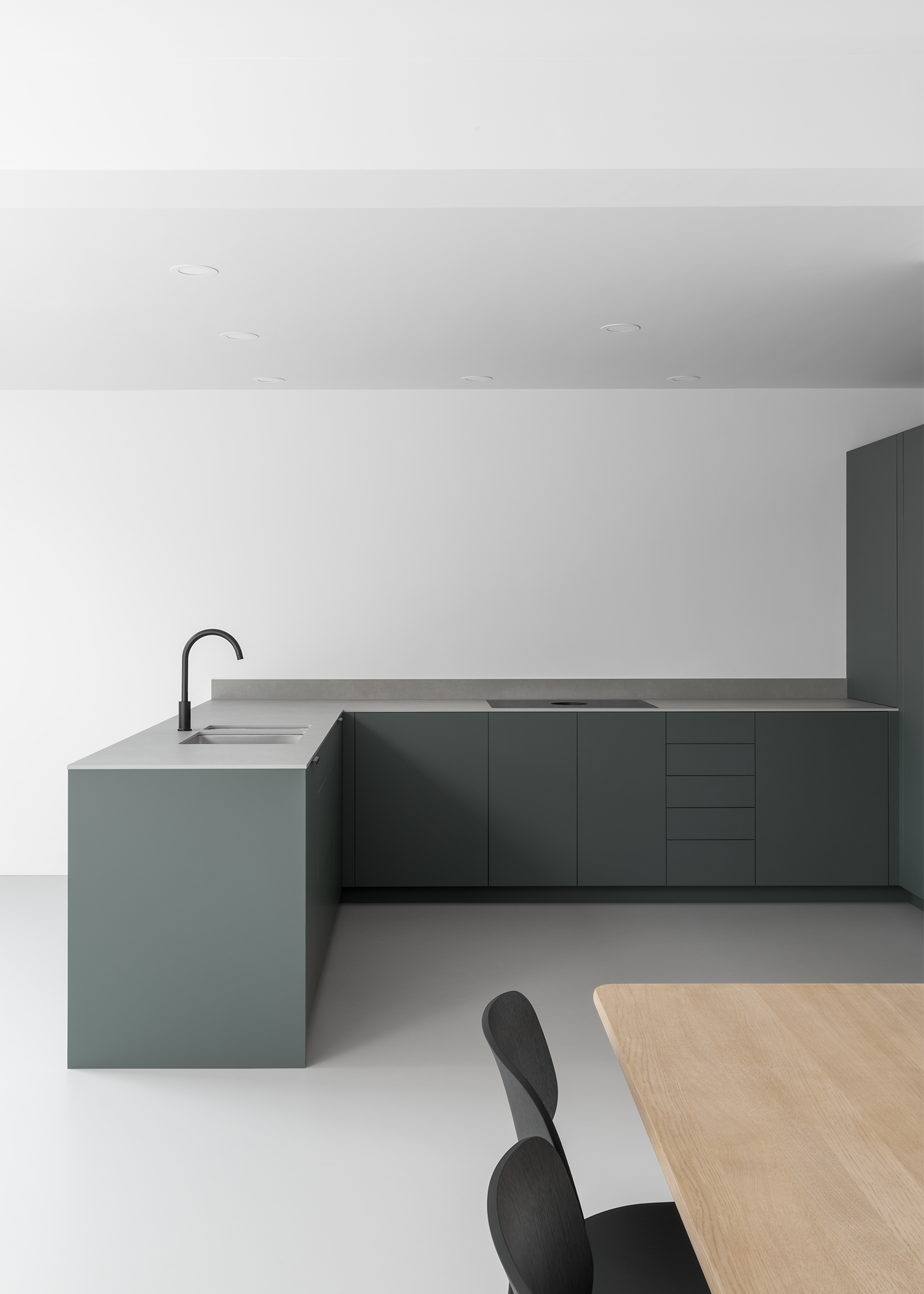Japanese style minimal modern white kitchen dining room with grey resin floor. Dark green-grey cabinets, textured grey slim work surface, black tap, Danish oak dining table with modern black chairs.