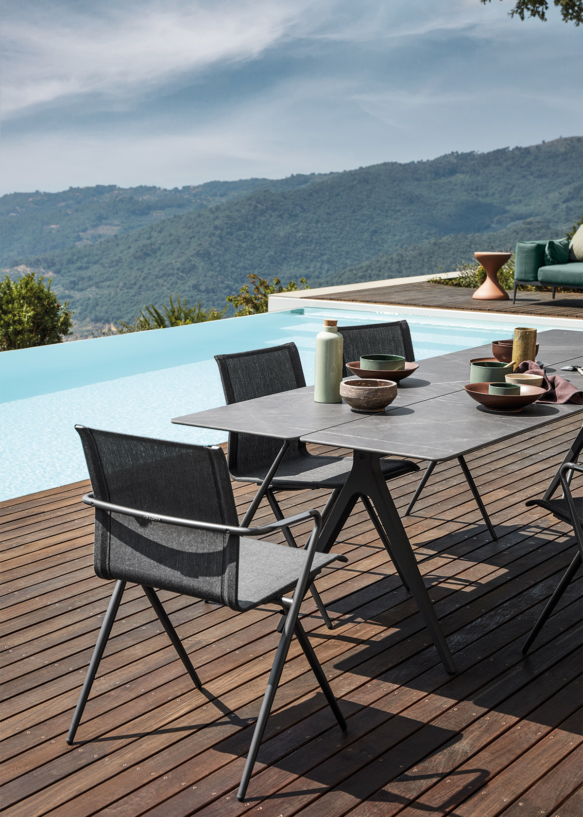 Split outdoor ceramic table and Ryder outdoor metal dining chair on wooden terrace next to modern infinity pool overlooking hills