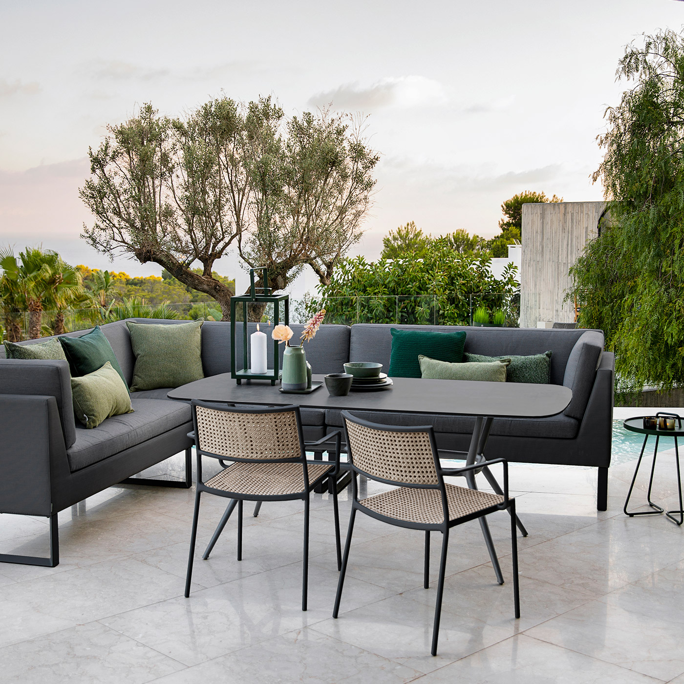 Less outdoor dining chair in modern cane with Joy outdoor dining table in grey aluminium and ceramic with a sofa on modern terrace with tropical plants overlooking the sea