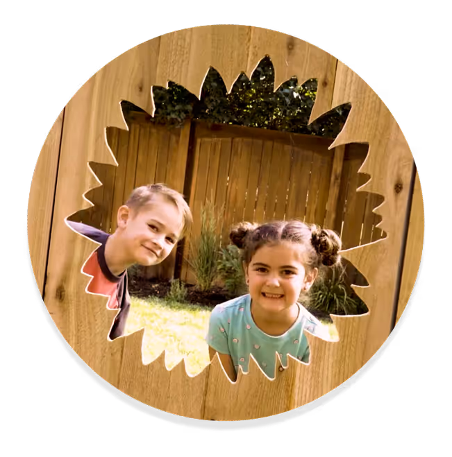 Two children smiling and looking through a sunflower-shaped cutout in a wooden fence in a backyard.