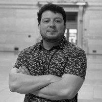 A gray-scale photo of Joaquin wearing a collared button-down t-shirt smiling with his arms crossed.