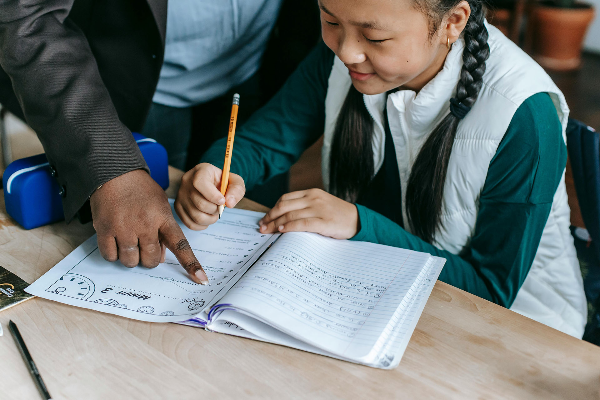 Teacher guiding an elementary student as she writes in a workbook at a classroom desk.