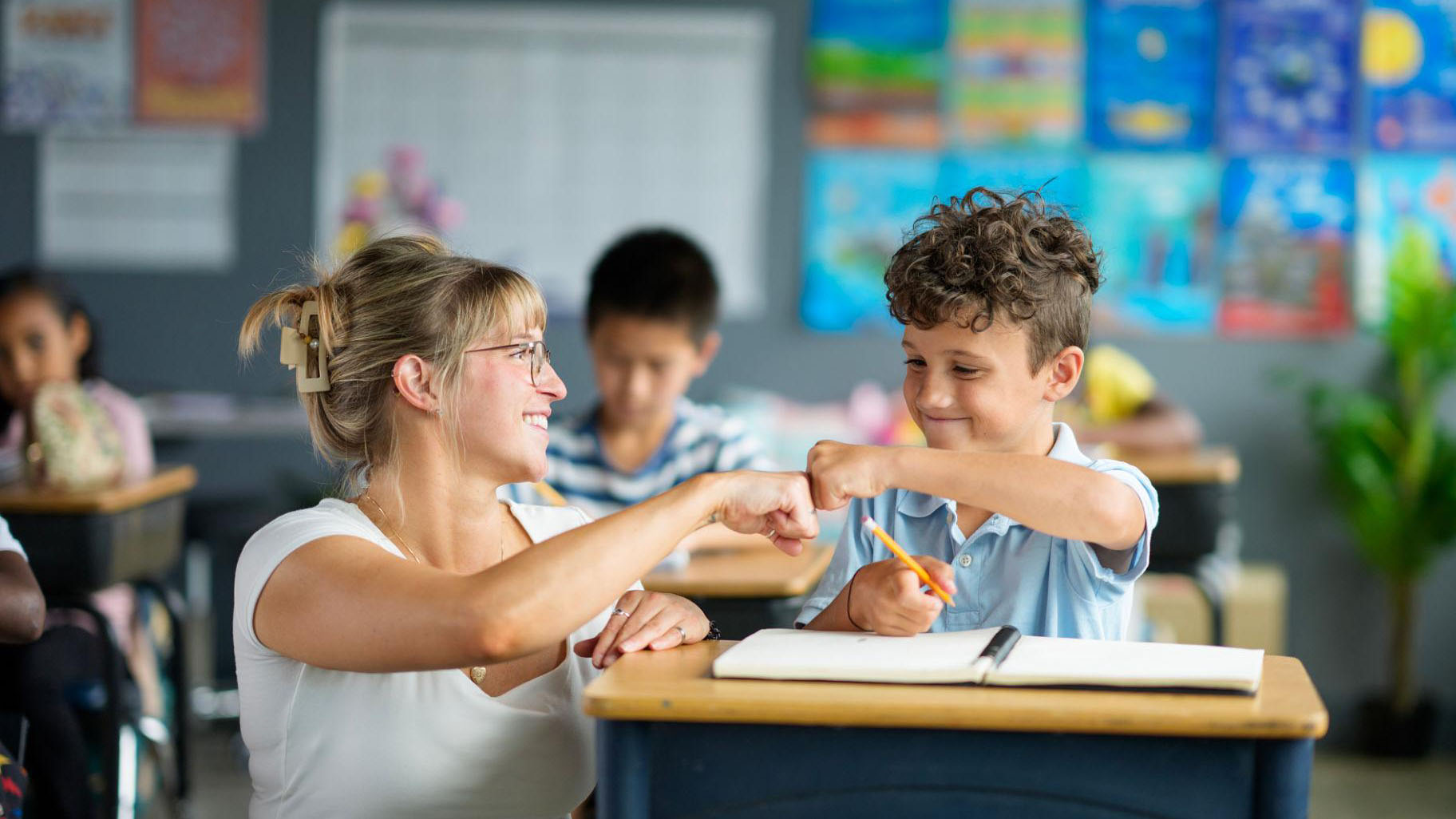 Teacher and student fist bumping at a desk in a classroom during a learning activity.