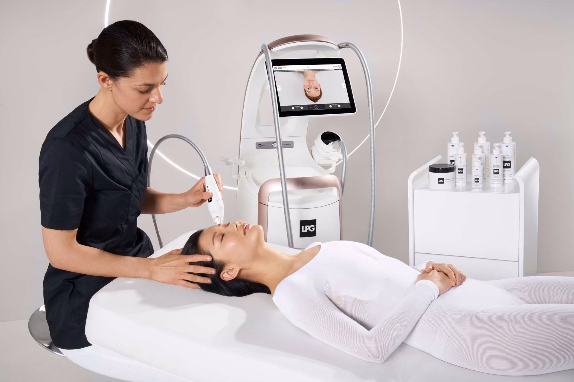 Woman in black applies facial treatment with device on a woman lying on a bed wearing white suit.