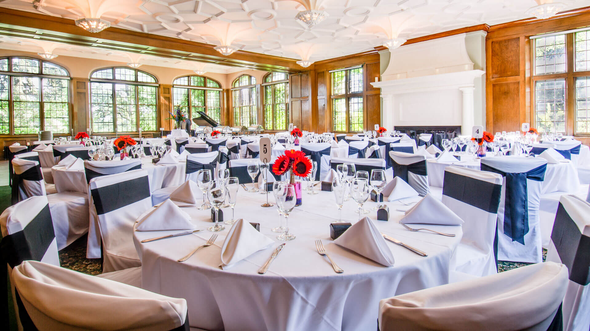 Elegant banquet room with round tables set for an event, white tablecloths, black chair sashes, and red floral centerpieces.