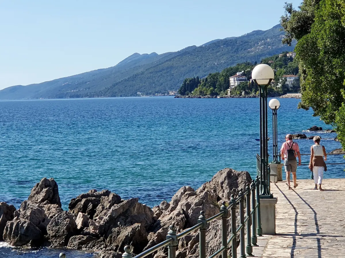 Coppia passeggia lungo il lungomare con vista sul mare blu e montagne, paesaggio rilassante e naturale