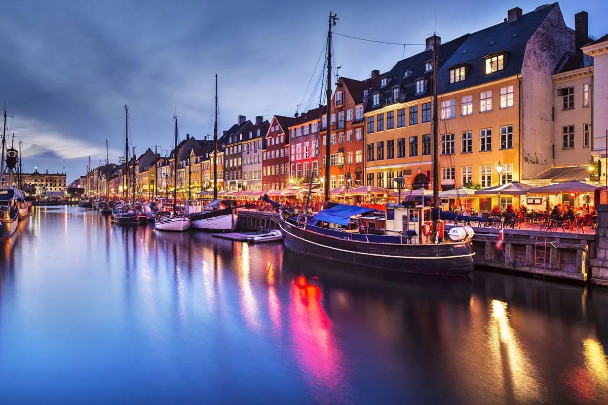 Vista serale di Nyhavn a Copenaghen, con barche attraccate, case colorate illuminate e riflessi sull’acqua.