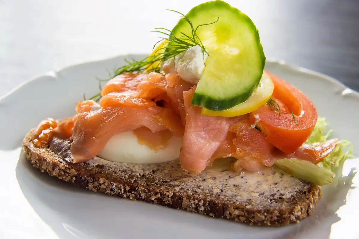 Smørrebrød danese con salmone affumicato, uovo sodo, cetriolo e pane di segale servito su piatto bianco.