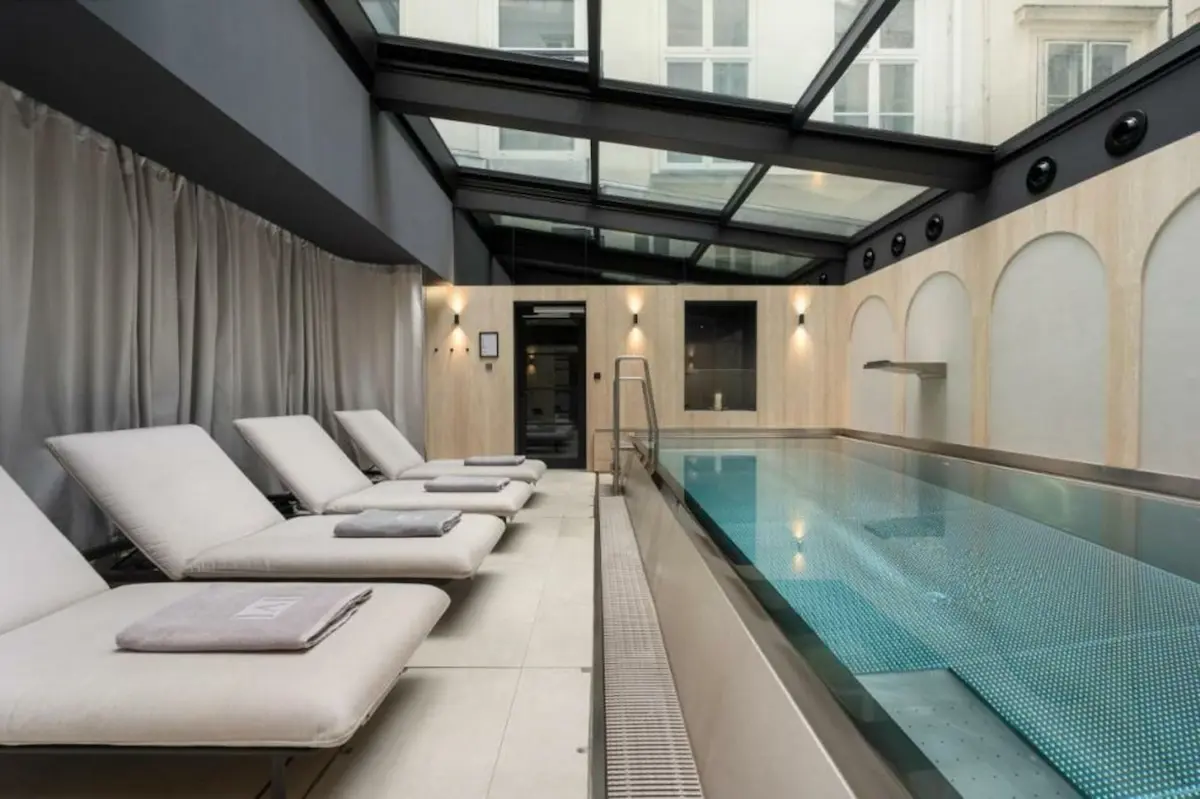 Piscina interna con lettini relax nella spa dell’hotel The Ring a Vienna, illuminata da luce naturale attraverso il soffitto vetrato