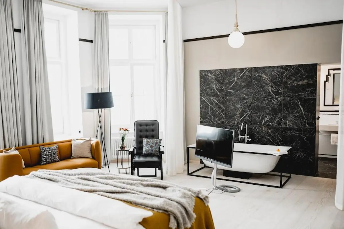 Suite di design dell’hotel Altstadt Vienna con letto, divano senape, poltrona, vasca freestanding e parete in marmo scuro
